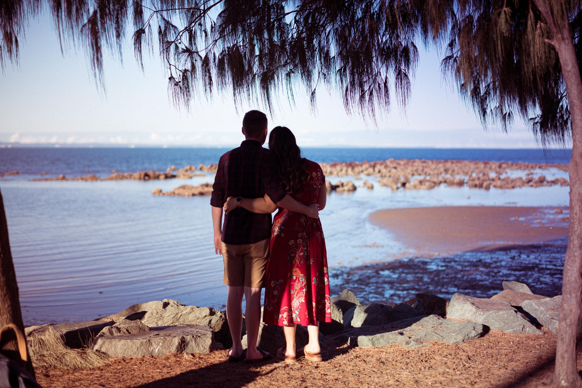 The couple stands arm in arm, looking out over the water at Redcliffe, under a tree.