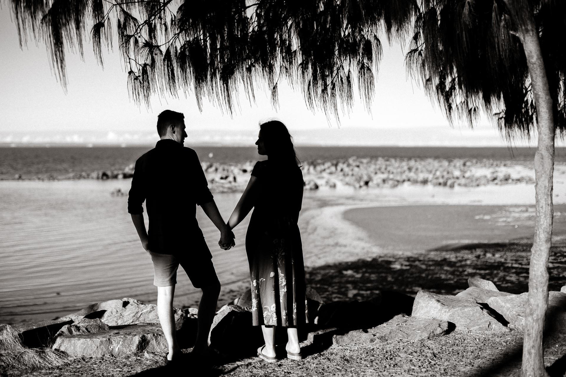 The couple holds hands, silhouetted against the sea at Redcliffe, under a tree.