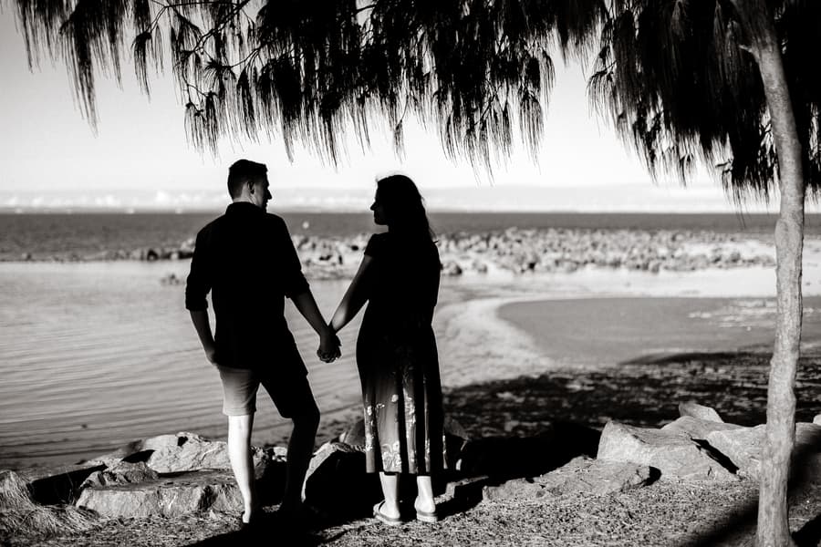The couple holds hands, silhouetted against the sea at Redcliffe, under a tree.