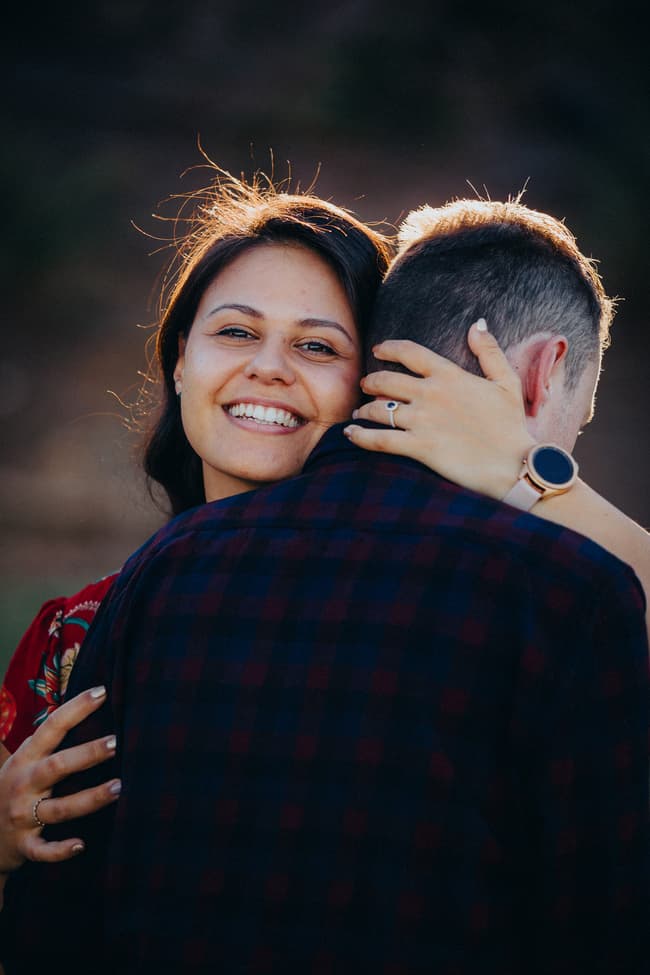 The couple embraces, one smiling brightly over the other's shoulder in warm light.