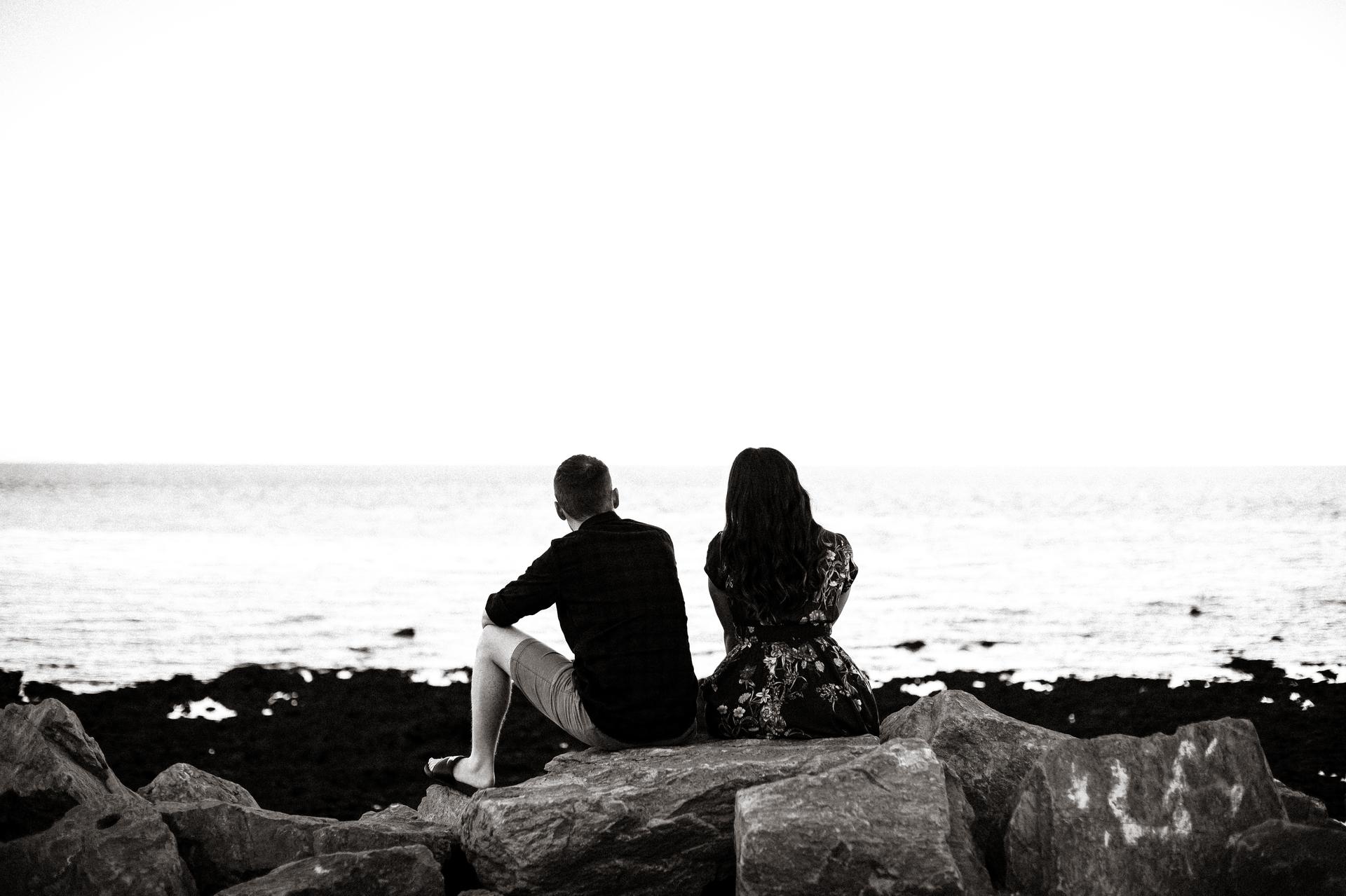 The couple sits on rocks at Redcliffe, looking out over the sea in a black and white photo.