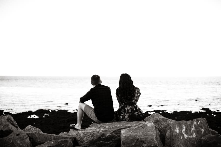 The couple sits on rocks at Redcliffe, looking out over the sea in a black and white photo.