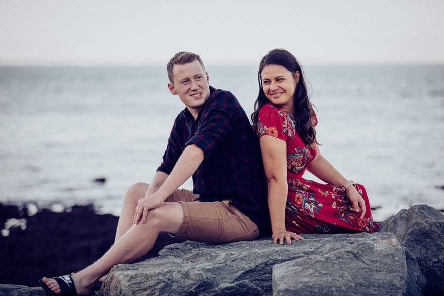 The couple sits back to back on rocks at Redcliffe, smiling with the sea in the background.