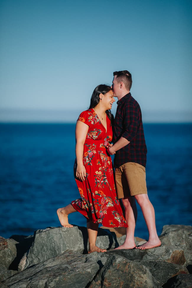 Couple standing on rocks by the sea at Redcliffe, with the man gently kissing the woman's forehead under a clear blue sky.