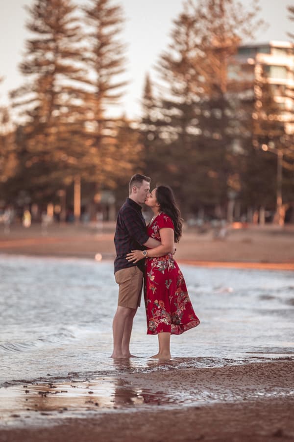 Couple standing barefoot on the beach at Redcliffe, embracing and gazing into each other's eyes with tall trees in the background.