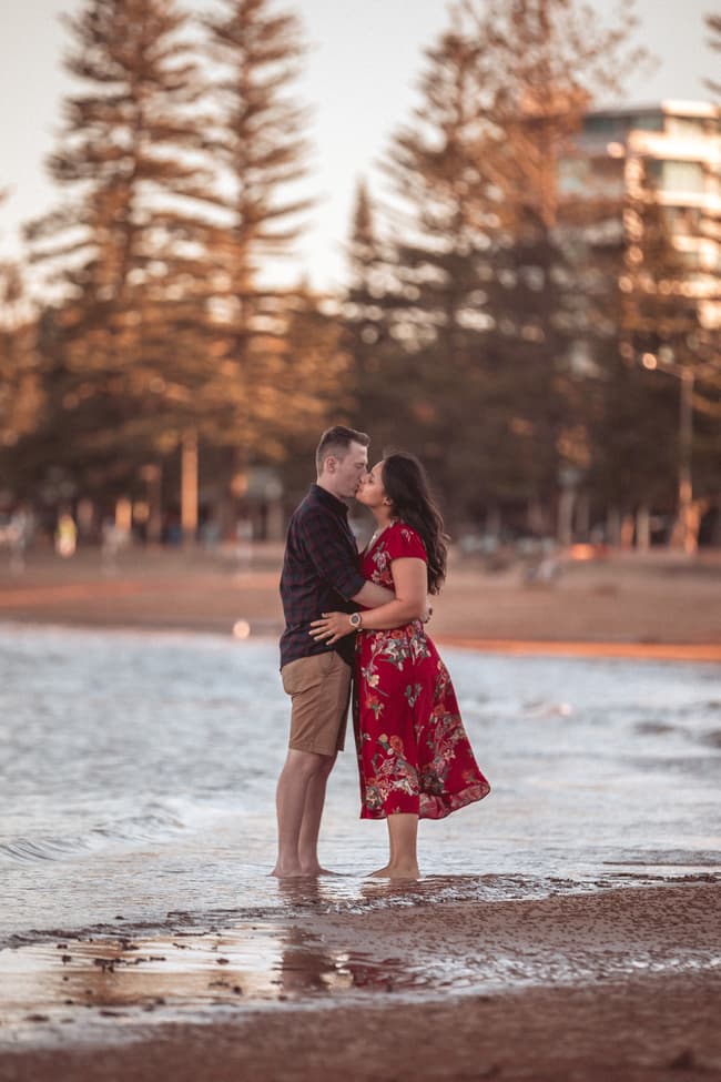 Couple standing barefoot on the beach at Redcliffe, embracing and gazing into each other's eyes with tall trees in the background.