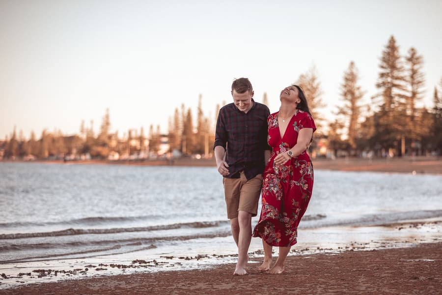 Couple walking barefoot along the shoreline at Redcliffe, laughing together with trees and water in the background.
