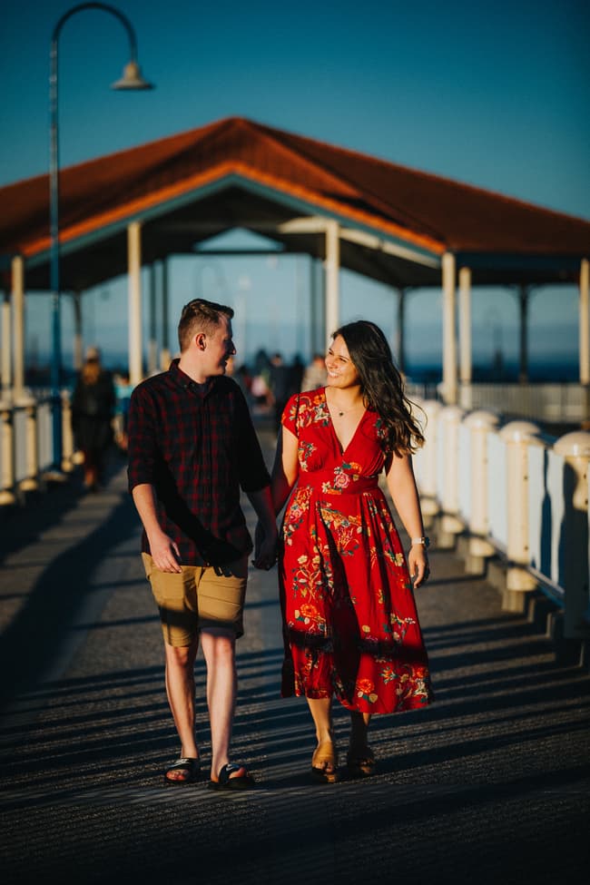 Couple walking hand in hand on a pier at Redcliffe, both smiling warmly at each other under a sunny sky.