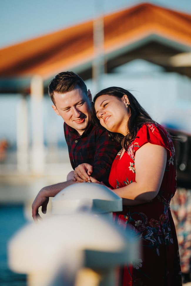 Couple leaning on a railing at Redcliffe Pier, the woman resting her head on the man's shoulder, both smiling softly.