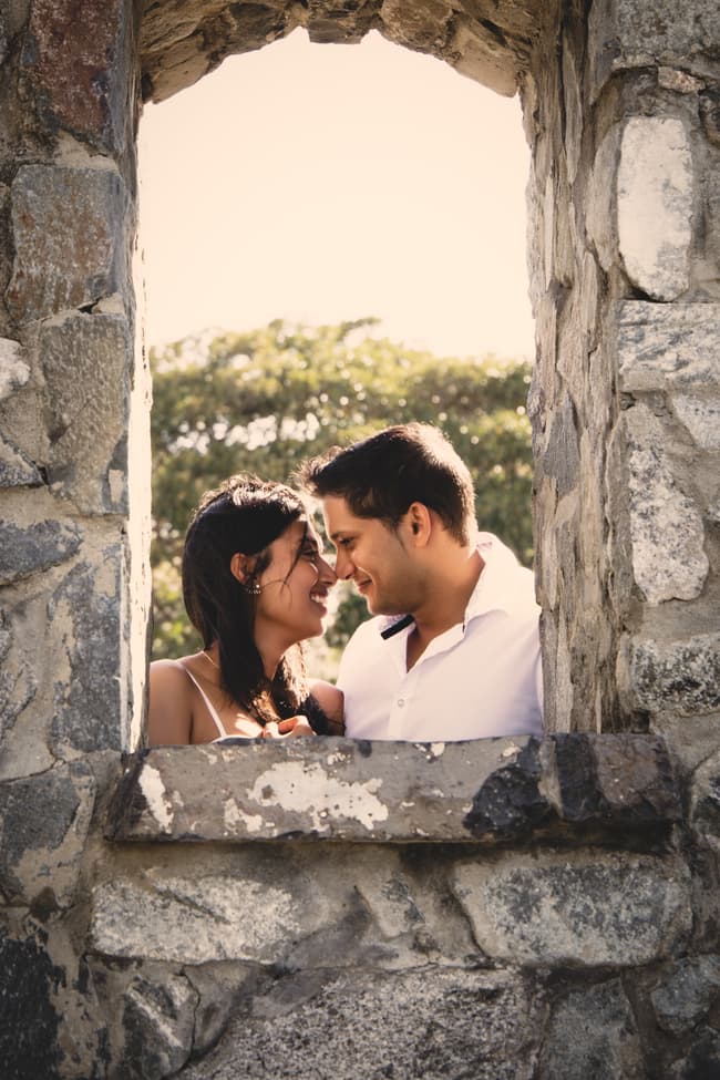 The couple smiling at each other through a stone window with greenery in the background.