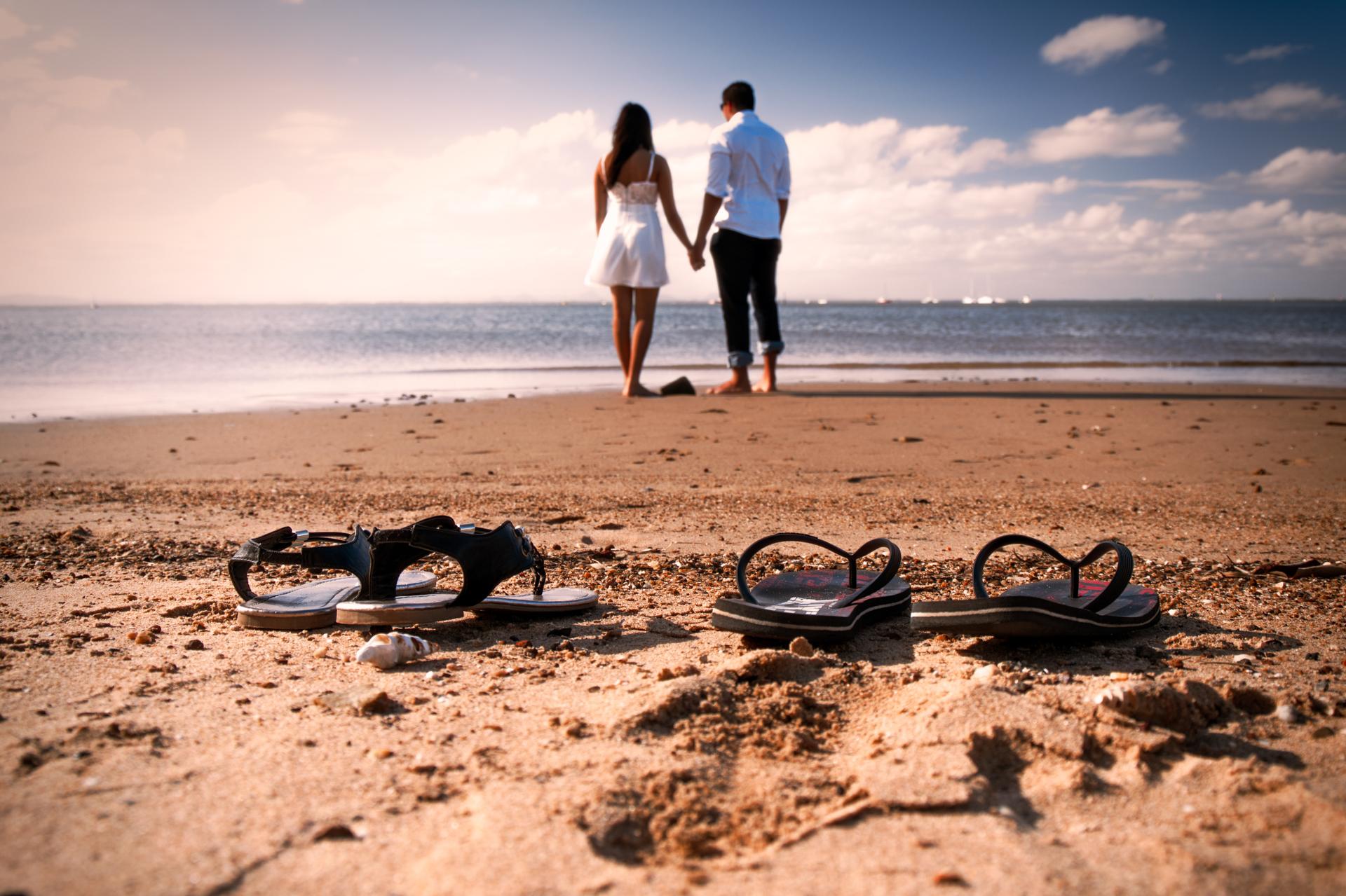 A couple walks hand in hand on Scarborough Beach, with sandals left on the sand in the foreground.
