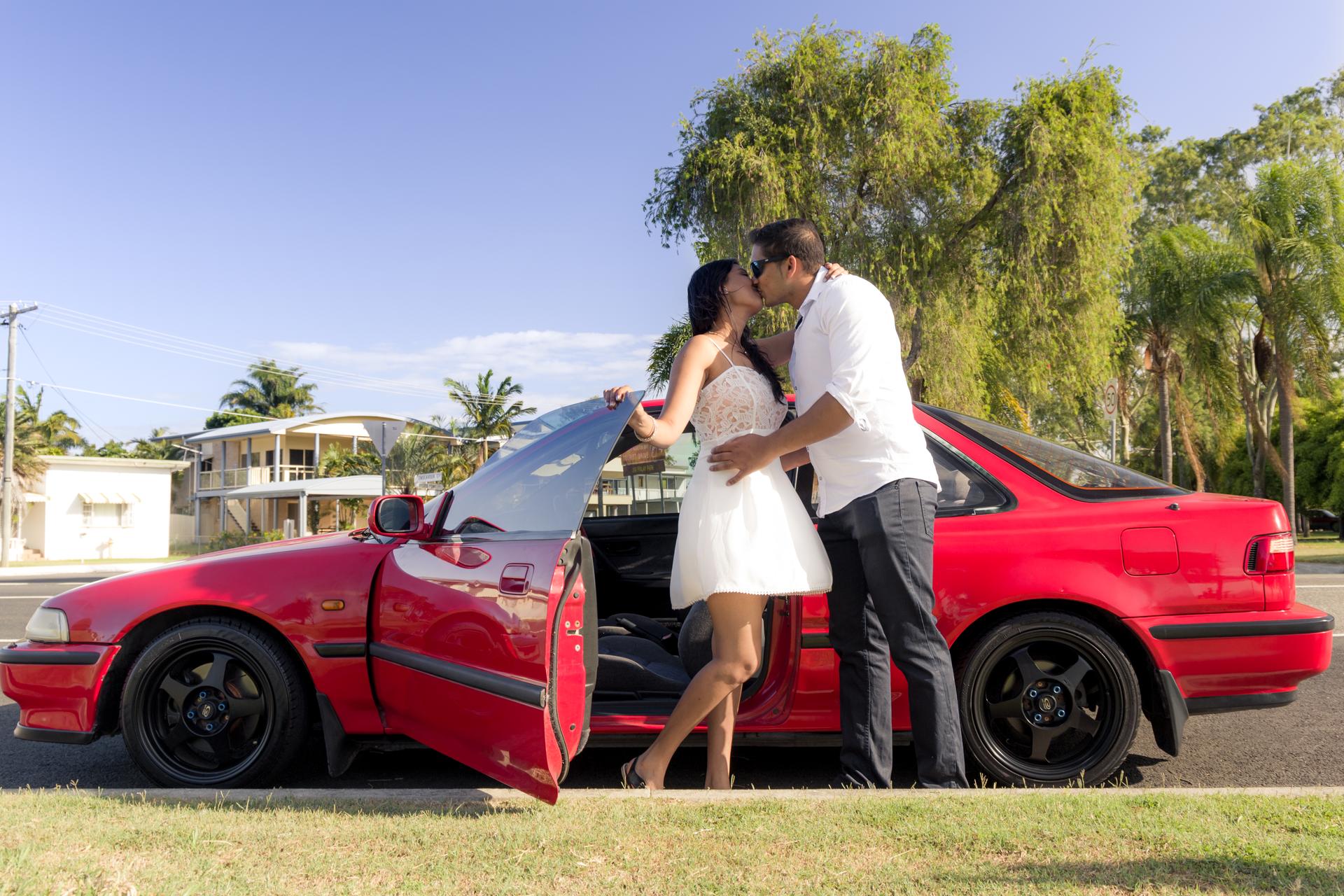 A couple stands beside a red car at Scarborough Beach, sharing a kiss under a clear blue sky.