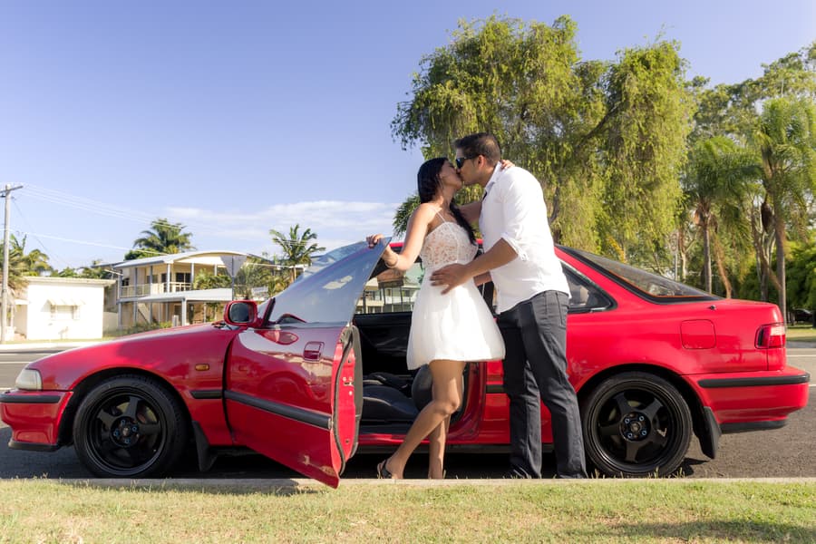 A couple stands beside a red car at Scarborough Beach, sharing a kiss under a clear blue sky.