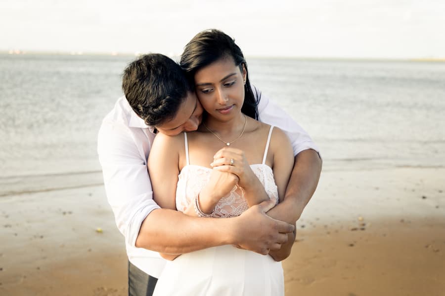 A couple stands on Scarborough Beach, with the person in front leaning back into the other's embrace.