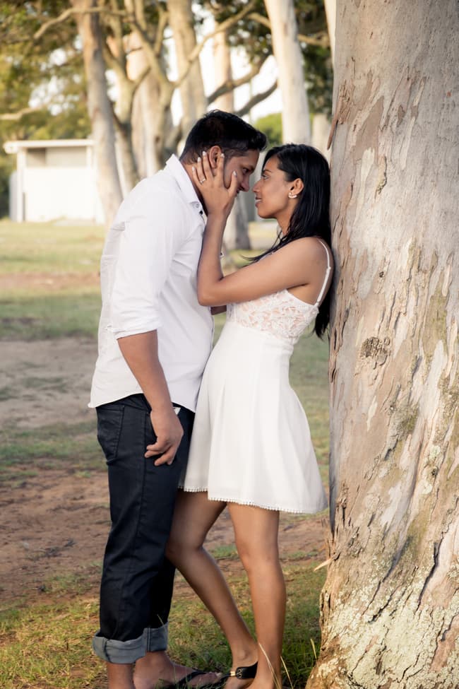 A couple stands close to a tree at Scarborough Beach, gazing into each other's eyes.