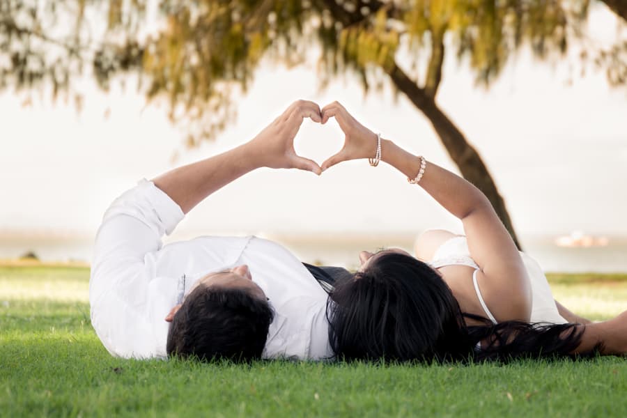 A couple lies on the grass at Scarborough Beach, forming a heart shape with their hands above them.