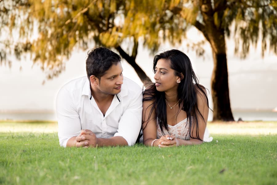 The couple lying on grass at Scarborough Beach, facing each other with trees in the background.