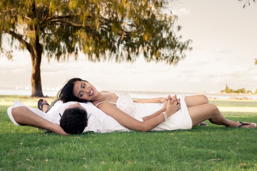 The couple lying on grass, one resting on the other's stomach, smiling under a tree at Scarborough Beach.