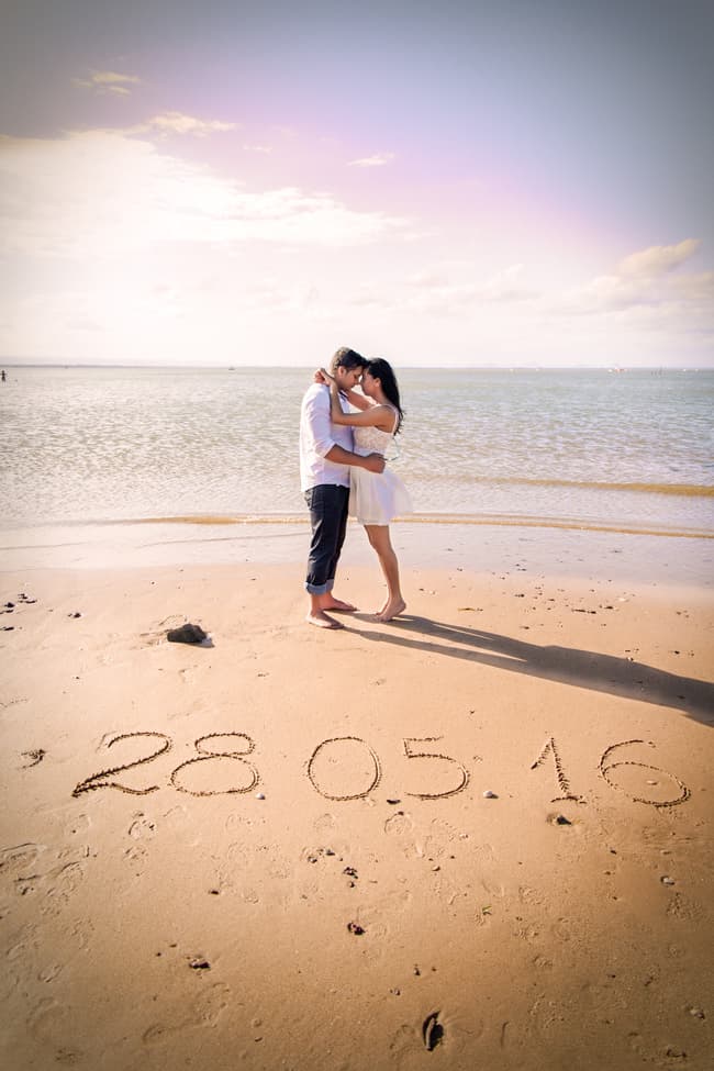 A couple embraces on Scarborough Beach with a date written in the sand in front of them.