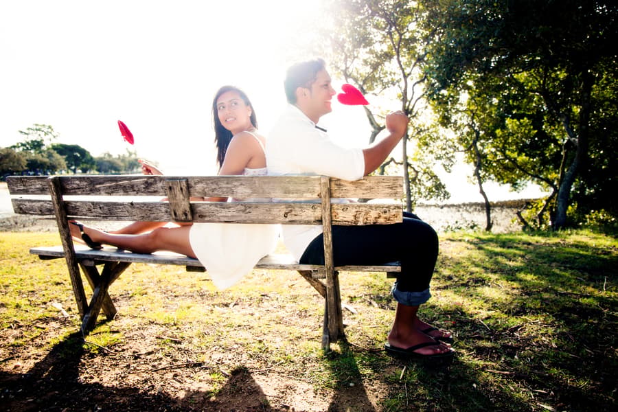A couple sits back-to-back on a bench at Scarborough Beach, each holding a red heart.