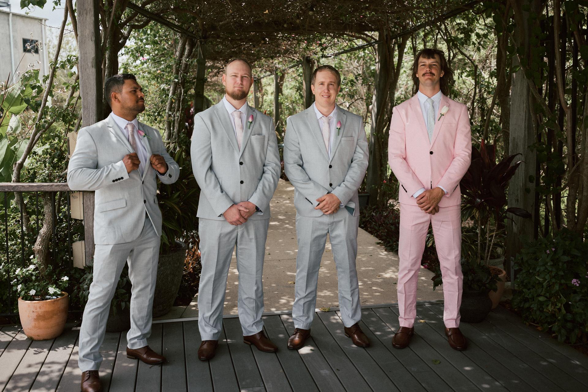Four groomsmen stand under a wooden pergola with greenery at Eatons Hill Hotel — Lakeside, three wearing light grey suits with pink ties and one wearing a pink suit with a white shirt and tie.