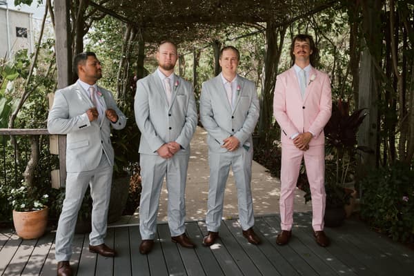 Four groomsmen stand under a wooden pergola with greenery at Eatons Hill Hotel — Lakeside, three wearing light grey suits with pink ties and one wearing a pink suit with a white shirt and tie.