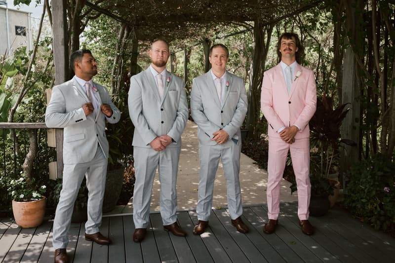 Four groomsmen stand under a wooden pergola with greenery at Eatons Hill Hotel — Lakeside, three wearing light grey suits with pink ties and one wearing a pink suit with a white shirt and tie.