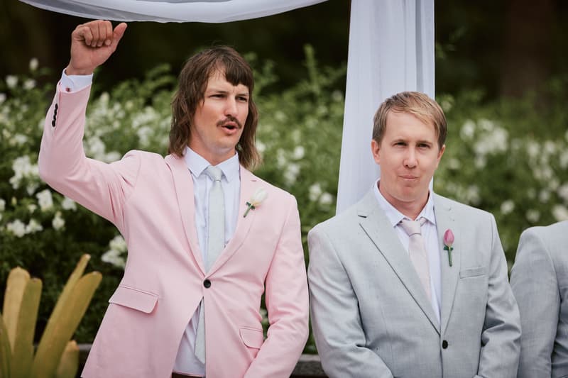 Jake, the groom, in a light pink suit raises his fist while standing next to a groomsman in a light grey suit with a pink boutonniere at the ceremony stage at Eatons Hill Hotel — Lakeside.