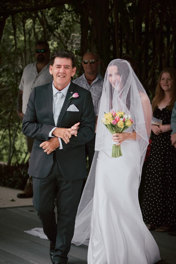 The bride Olivia, holding a bouquet of yellow and pink flowers, walks arm-in-arm with an older man, likely her father, at the Eatons Hill Hotel — Lakeside ceremony stage. Guests stand in the background watching.