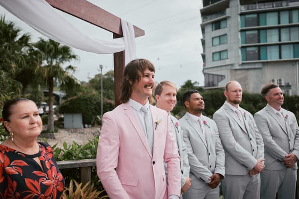 Jake the groom in a pink suit stands with his groomsmen dressed in light grey suits with pink boutonnieres at the ceremony stage of Eatons Hill Hotel — Lakeside. A woman in a floral dress stands nearby.