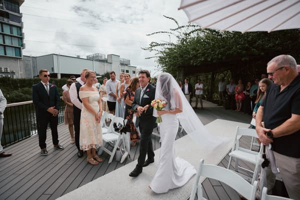 The bride Olivia, holding a bouquet, is walking down the aisle escorted by an older man, likely her father, at the ceremony stage at Eatons Hill Hotel — Lakeside. Guests stand on either side of the aisle watching the procession.