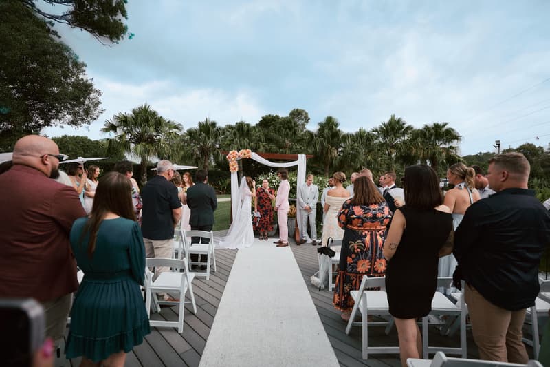 Olivia and Jake stand under a floral arch at the ceremony stage at Eatons Hill Hotel — Lakeside, surrounded by guests seated on white chairs along the aisle.