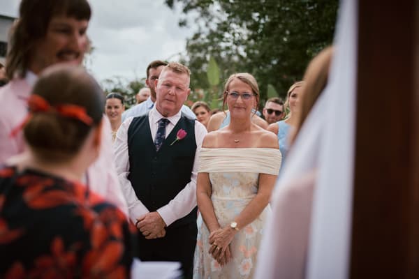 The bride's parents stand together among guests at the ceremony stage at Eatons Hill Hotel — Lakeside, with bridesmaids visible in the background.