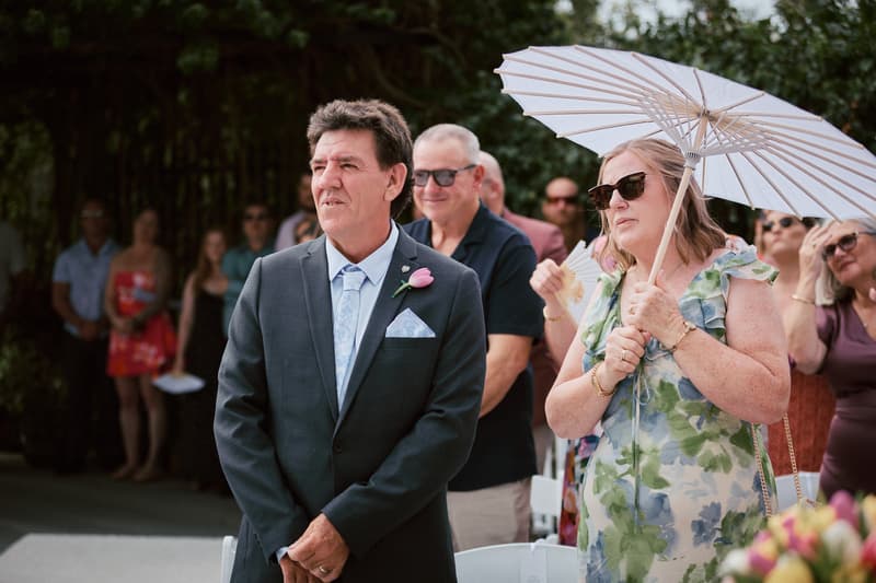 Guests stand and watch during the ceremony at Eatons Hill Hotel — Lakeside, including a man in a suit with a pink boutonniere and a woman holding a white parasol wearing a floral dress.