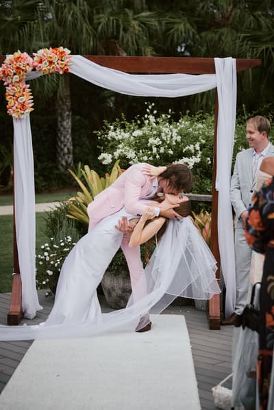 Olivia and Jake share a kiss under a floral wedding arch at Eatons Hill Hotel — Lakeside during their ceremony, with a guest watching nearby.