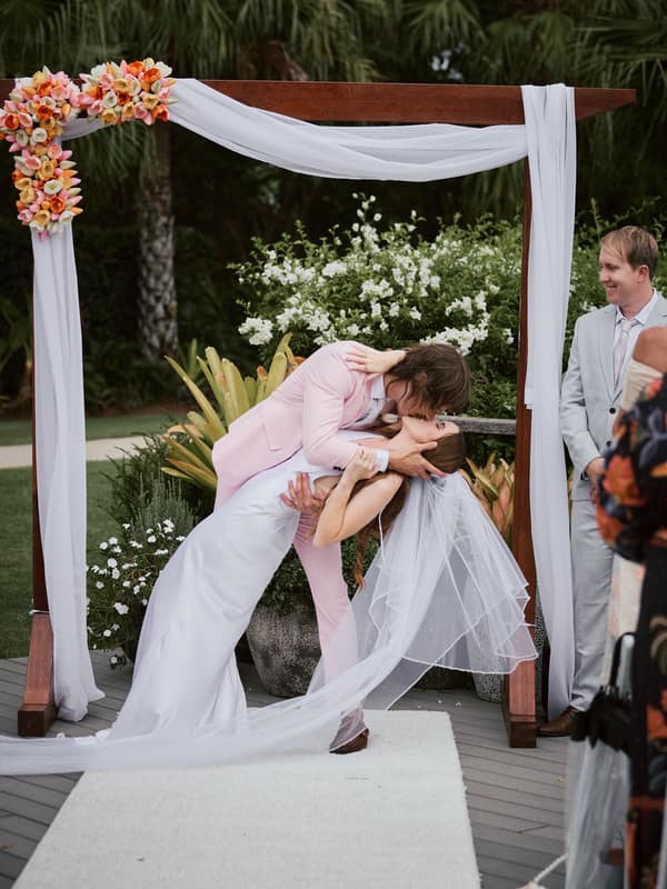 Olivia and Jake share a kiss under a floral wedding arch at Eatons Hill Hotel — Lakeside during their ceremony, with a guest watching nearby.