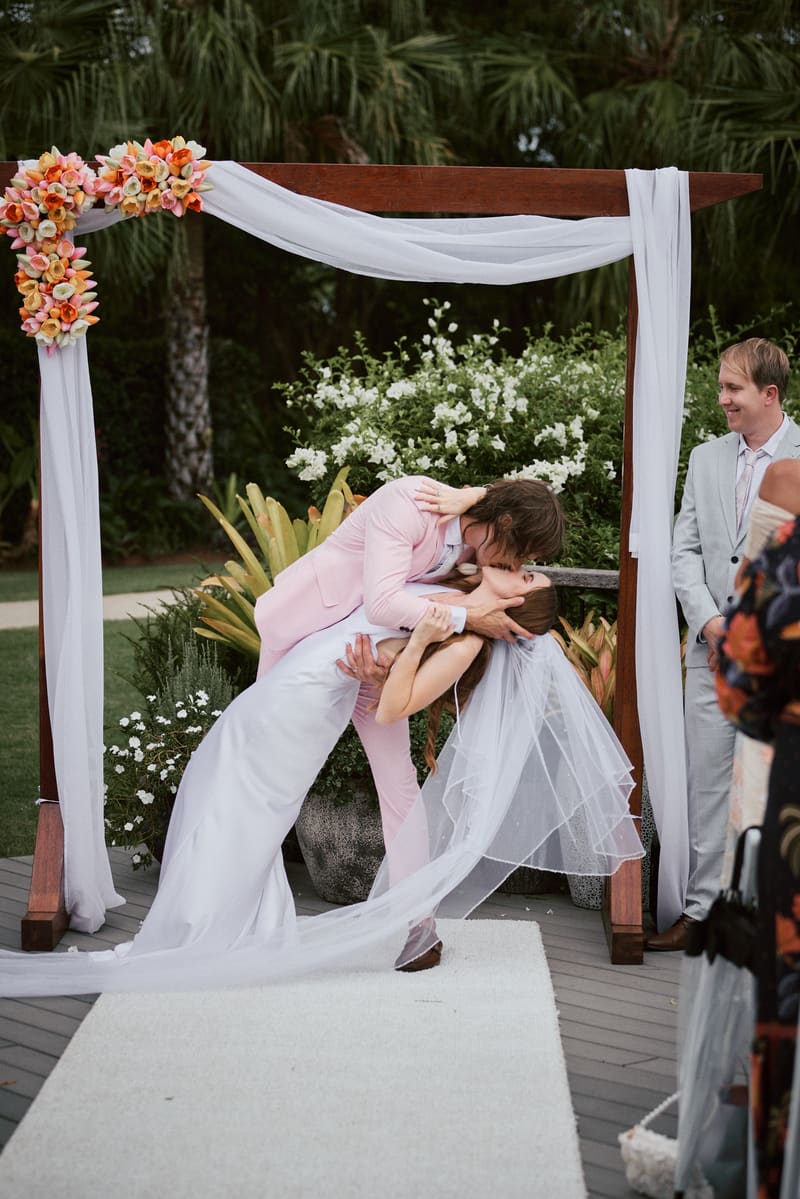 Olivia and Jake share a kiss under a floral wedding arch at Eatons Hill Hotel — Lakeside during their ceremony, with a guest watching nearby.