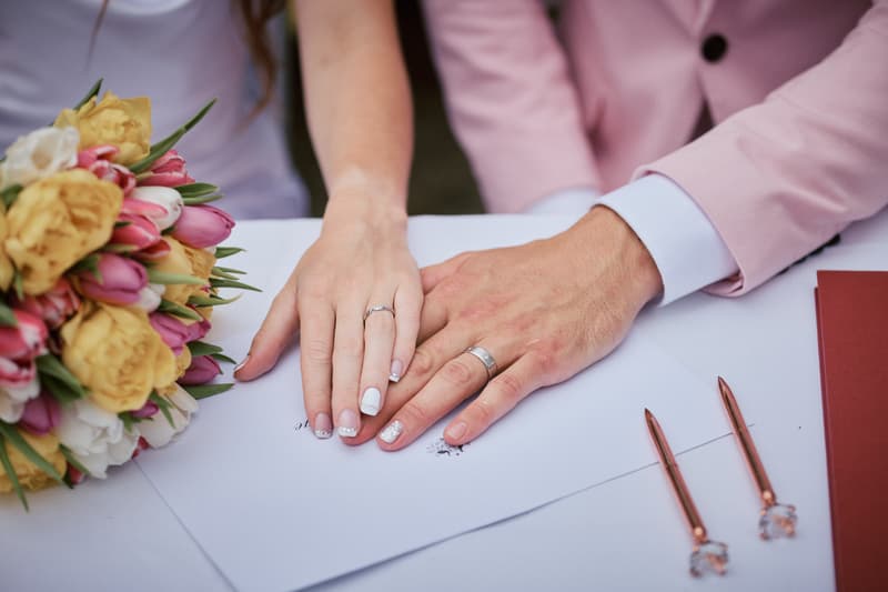 The bride Olivia and groom Jake place their hands together on a signed document at the ceremony stage at Eatons Hill Hotel — Lakeside, with a bouquet and two pens visible on the table.