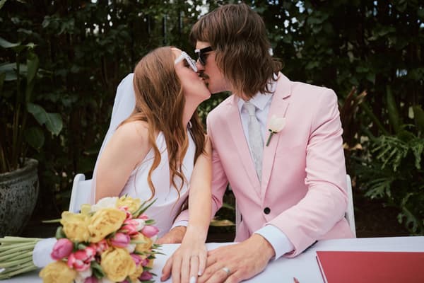 Olivia the bride and Jake the groom kiss while seated at a table with a bouquet of yellow and pink flowers at Eatons Hill Hotel — Lakeside.