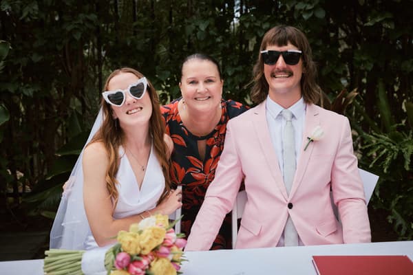 Bride Olivia and groom Jake, both wearing sunglasses, sit at a table with a woman standing behind them at Eatons Hill Hotel — Lakeside. Olivia holds a bouquet of flowers on the table.