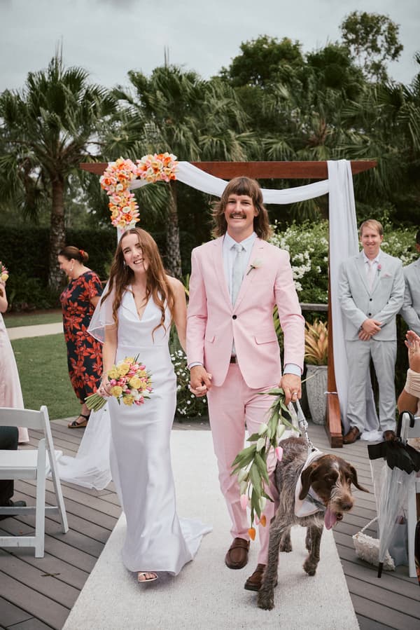Olivia the bride and Jake the groom walk down the aisle holding hands at Eatons Hill Hotel — Lakeside, accompanied by a dog wearing a floral garland, with a floral arch and guests in the background.