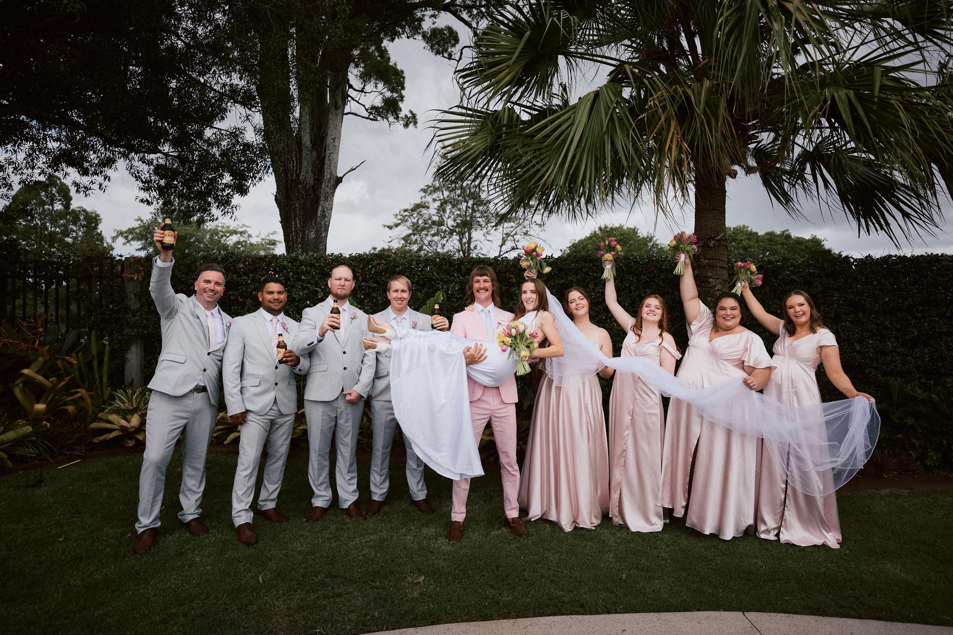 Jake in a pink suit holds Olivia in a white wedding dress, surrounded by groomsmen in light grey suits holding beers and bridesmaids in pale pink dresses holding bouquets at Eatons Hill Hotel outdoors.