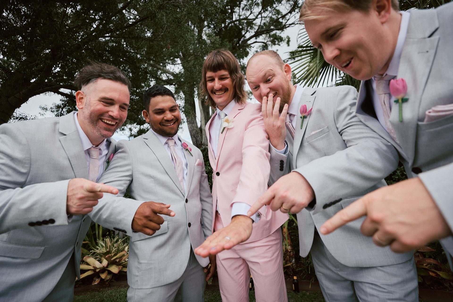 Jake in a pink suit shows his wedding ring to four groomsmen in grey suits with pink boutonnieres at Eatons Hill Hotel outdoors.