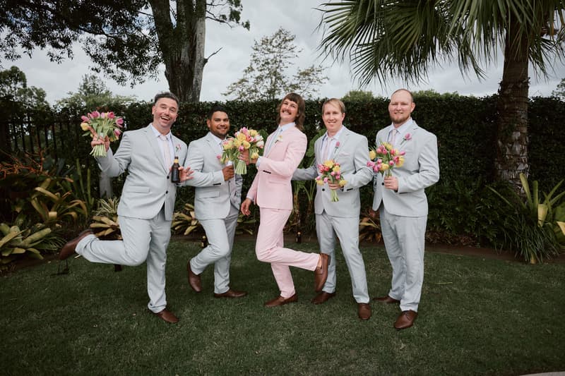 Jake in a pink suit and four groomsmen in light grey suits pose holding bouquets of flowers on the lawn at Eatons Hill Hotel.