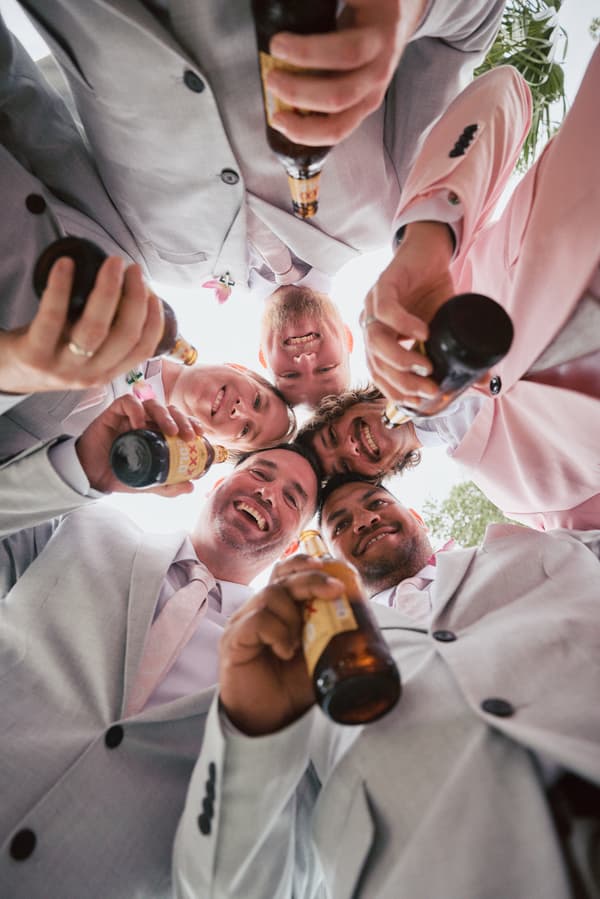 Five groomsmen in light grey suits with pink ties hold beer bottles and smile down at the camera in a group huddle.