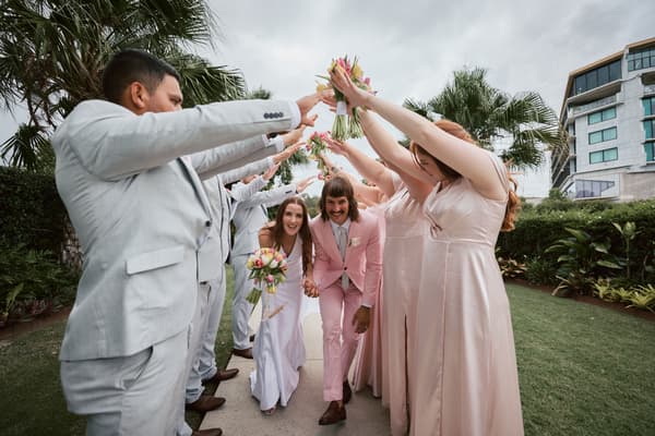 Olivia the bride and Jake the groom walk hand in hand under an arch formed by bridesmaids in blush dresses and groomsmen in light grey suits holding up bouquets and arms at Eatons Hill Hotel outdoor garden area.