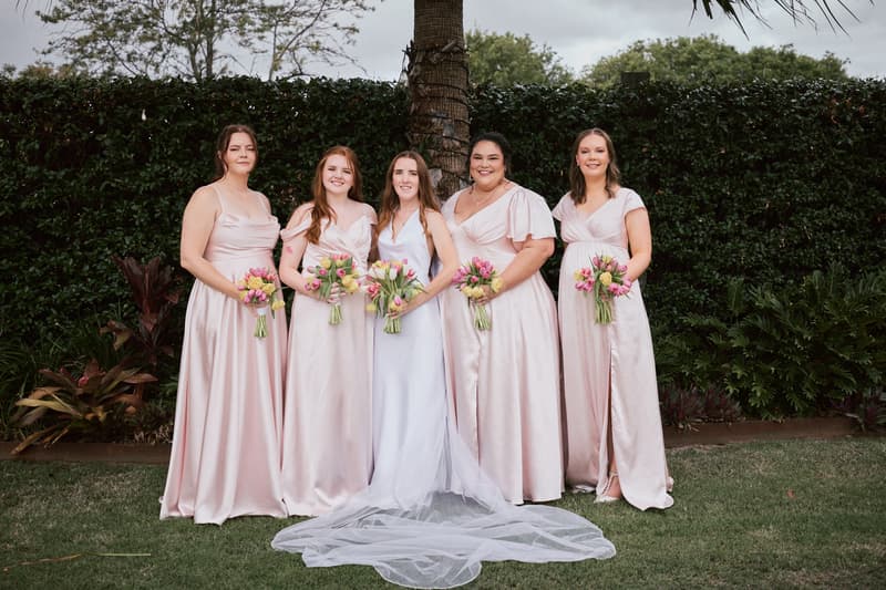 The bride Olivia stands with four bridesmaids in pale pink dresses holding bouquets in front of a hedge at Eatons Hill Hotel during the couple portraits stage.