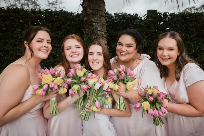 Five bridesmaids in light pink dresses holding bouquets of pink tulips and yellow flowers pose together outdoors at Eatons Hill Hotel.