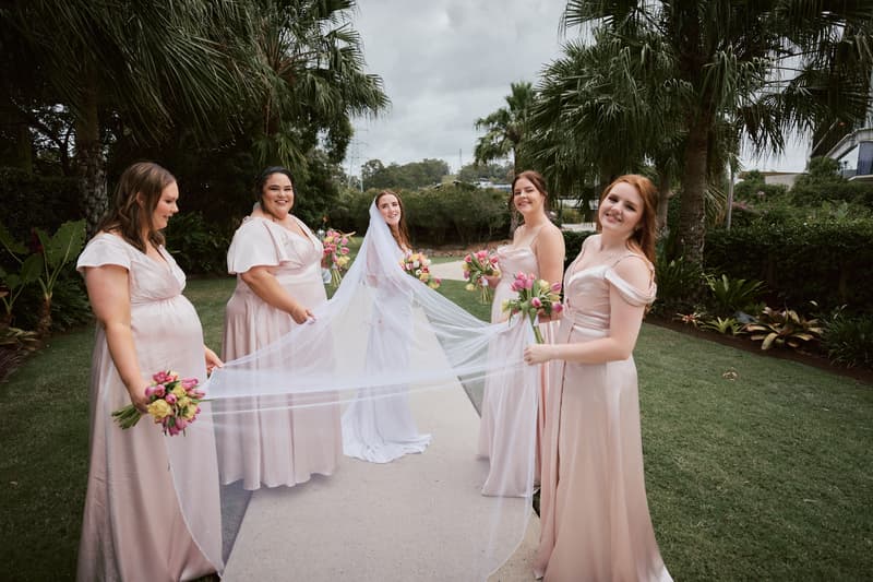 The bride Olivia stands on a garden pathway at Eatons Hill Hotel holding a bouquet, surrounded by four bridesmaids in matching blush pink dresses who are holding her veil and bouquets.