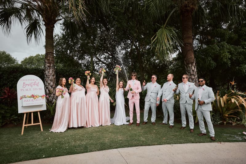 Olivia the bride in a white dress and Jake the groom in a pink suit stand with their bridal party at Eatons Hill Hotel. The bridesmaids wear matching light pink dresses and hold bouquets, while the groomsmen wear light grey suits. Jake is popping a champagne bottle, spraying the group. A wedding sign with their names and date is visible on the left.